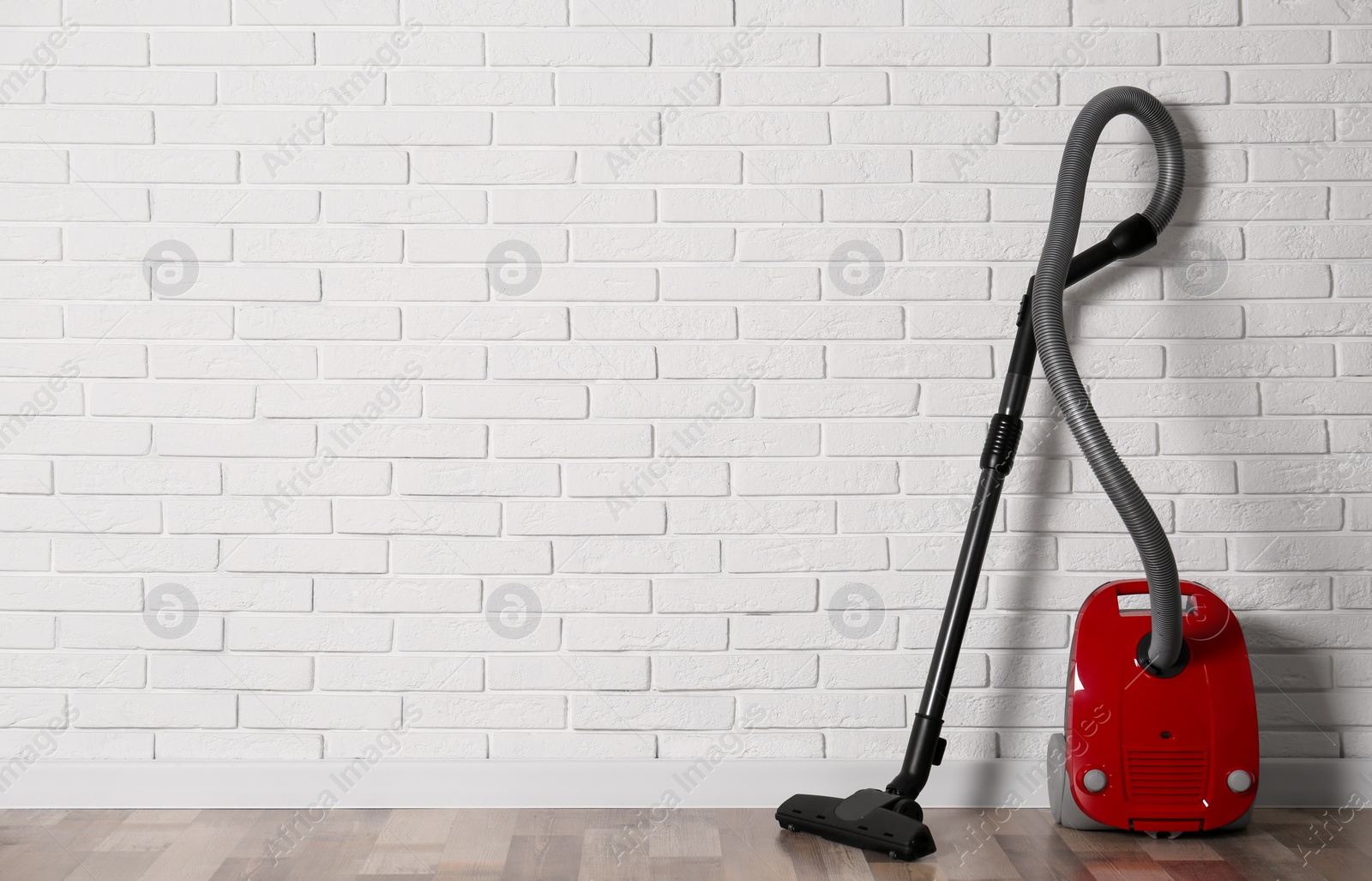 Photo of Modern red vacuum cleaner on wooden floor near white brick wall, space for text