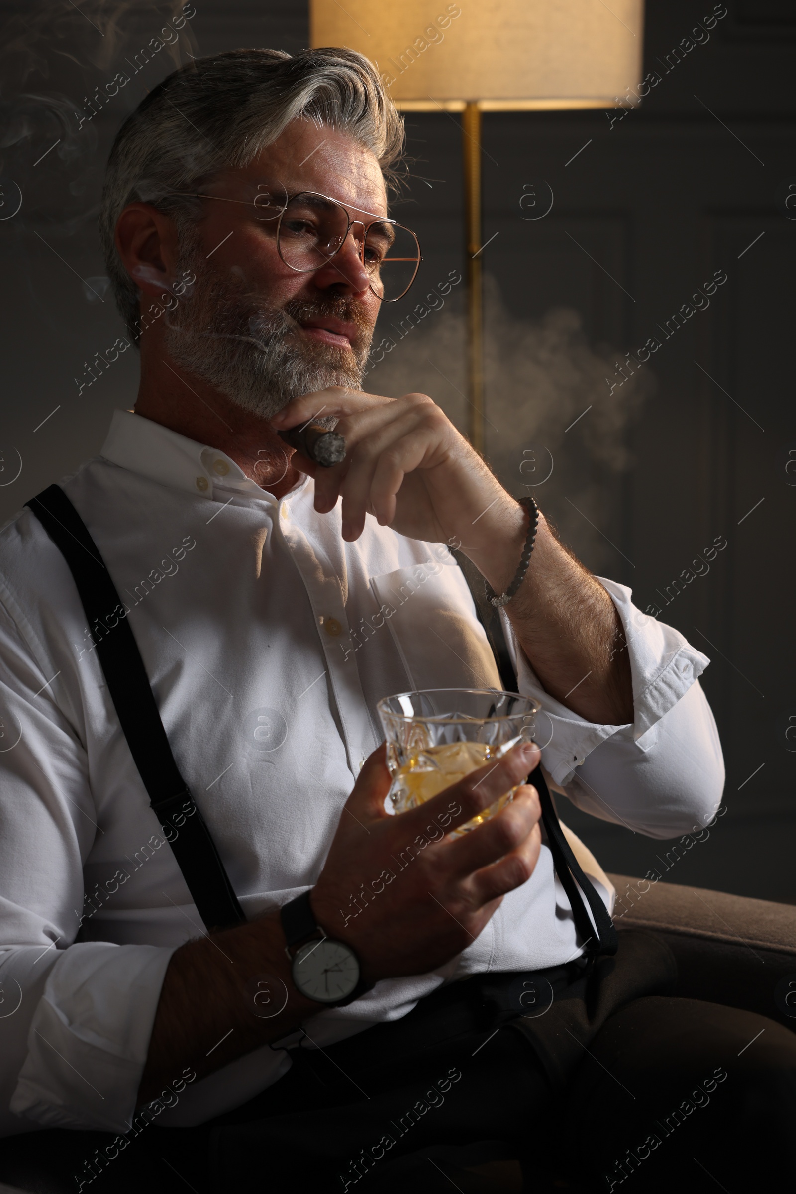 Photo of Bearded man with glass of whiskey smoking cigar in armchair indoors