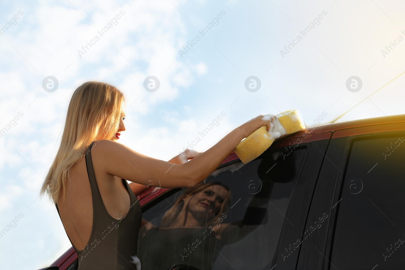 Photo of Young woman in swimsuits washing car with sponge outdoors