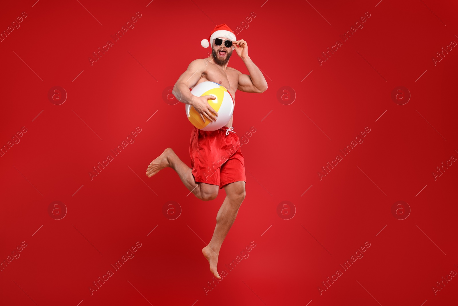 Photo of Muscular young man in Santa hat and sunglasses with ball jumping on red background