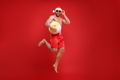 Muscular young man in Santa hat and sunglasses with ball jumping on red background