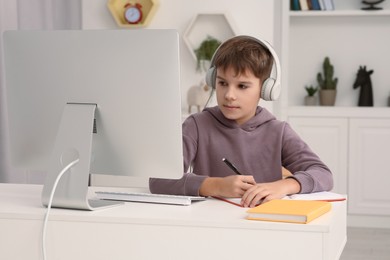 Photo of Boy writing in notepad while using computer and headphones at desk in room. Home workplace