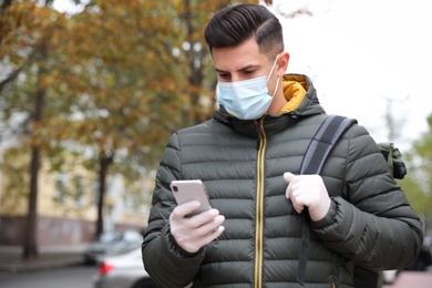 Man in medical face mask and gloves with smartphone walking outdoors. Personal protection during COVID-19 pandemic