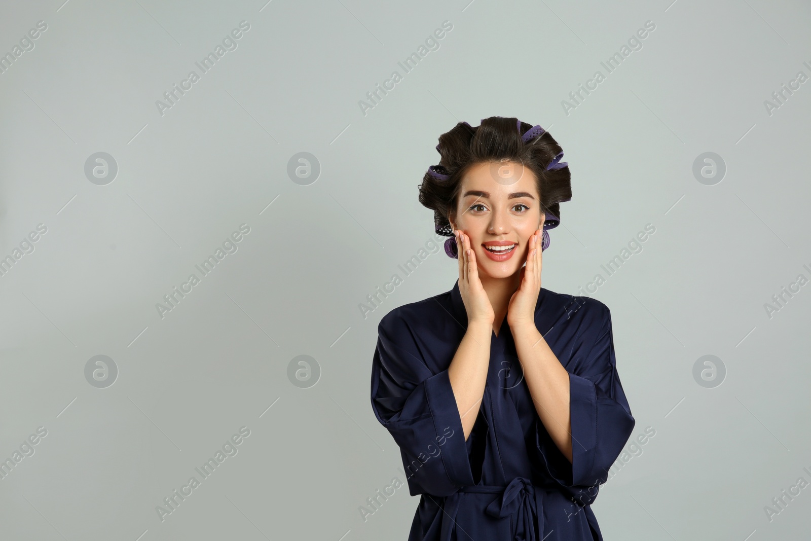 Photo of Happy young woman in silk bathrobe with hair curlers on grey background, space for text