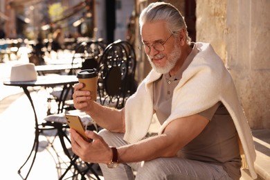 Photo of Handsome senior man sitting on doorstep, using smartphone and drinking coffee outdoors, space for text