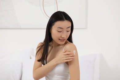 Beautiful young Asian woman applying body cream on shoulder in bedroom