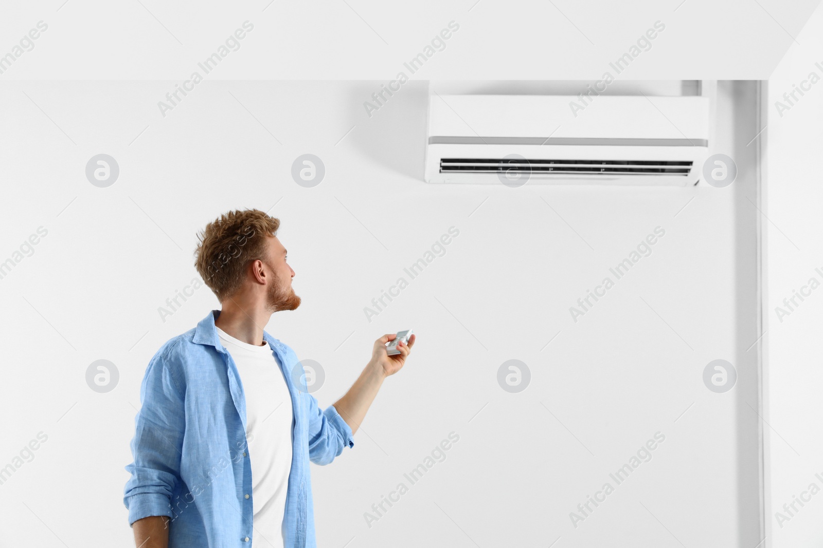 Photo of Young man operating air conditioner with remote control indoors. Space for text