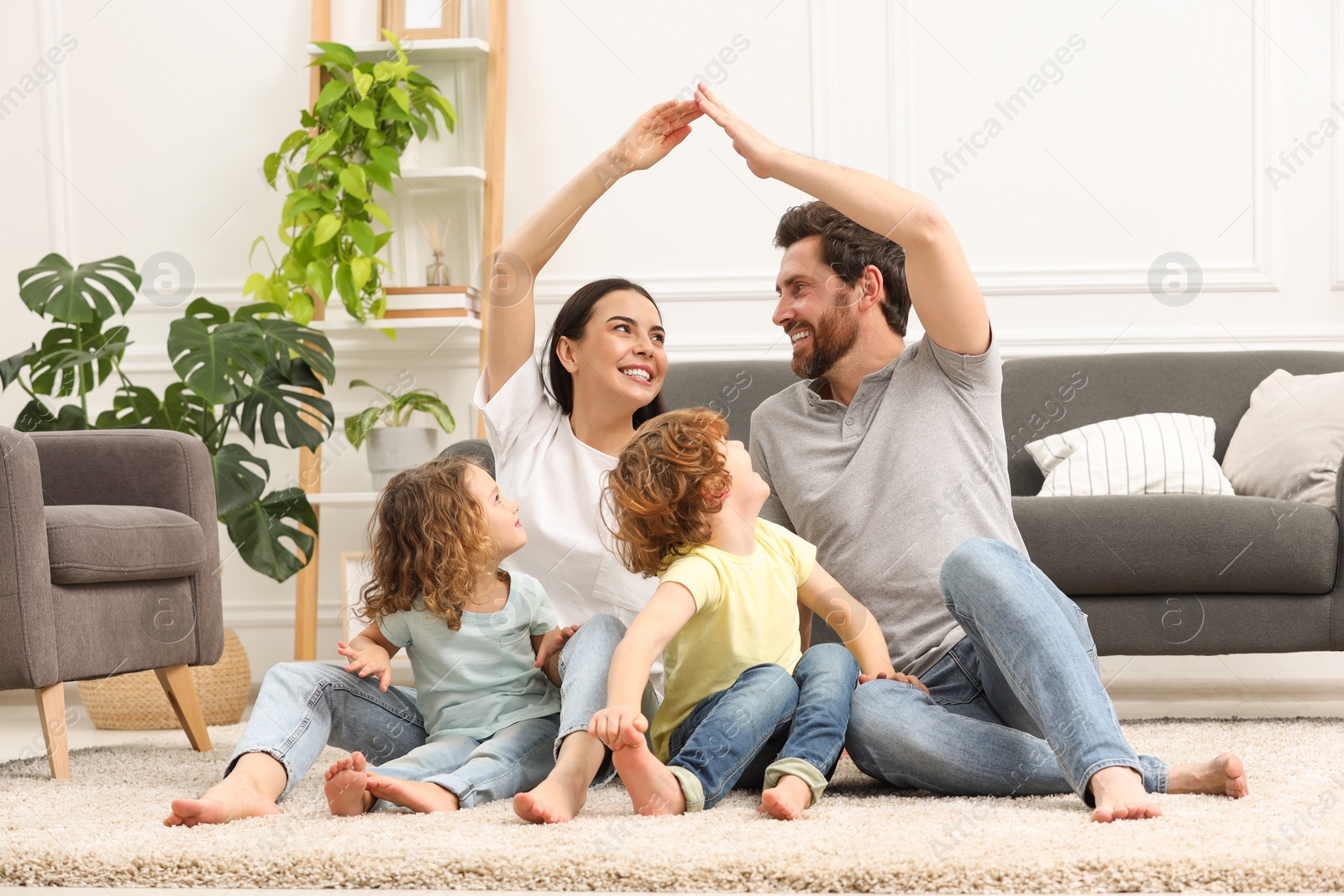 Photo of Family housing concept. Happy woman and her husband forming roof with their hands while sitting with kids on floor at home