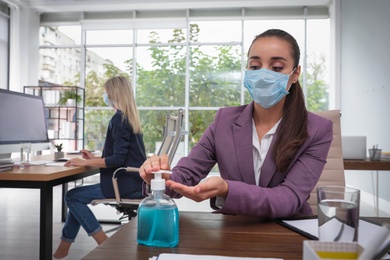 Photo of Office employee in mask applying hand sanitizer at workplace