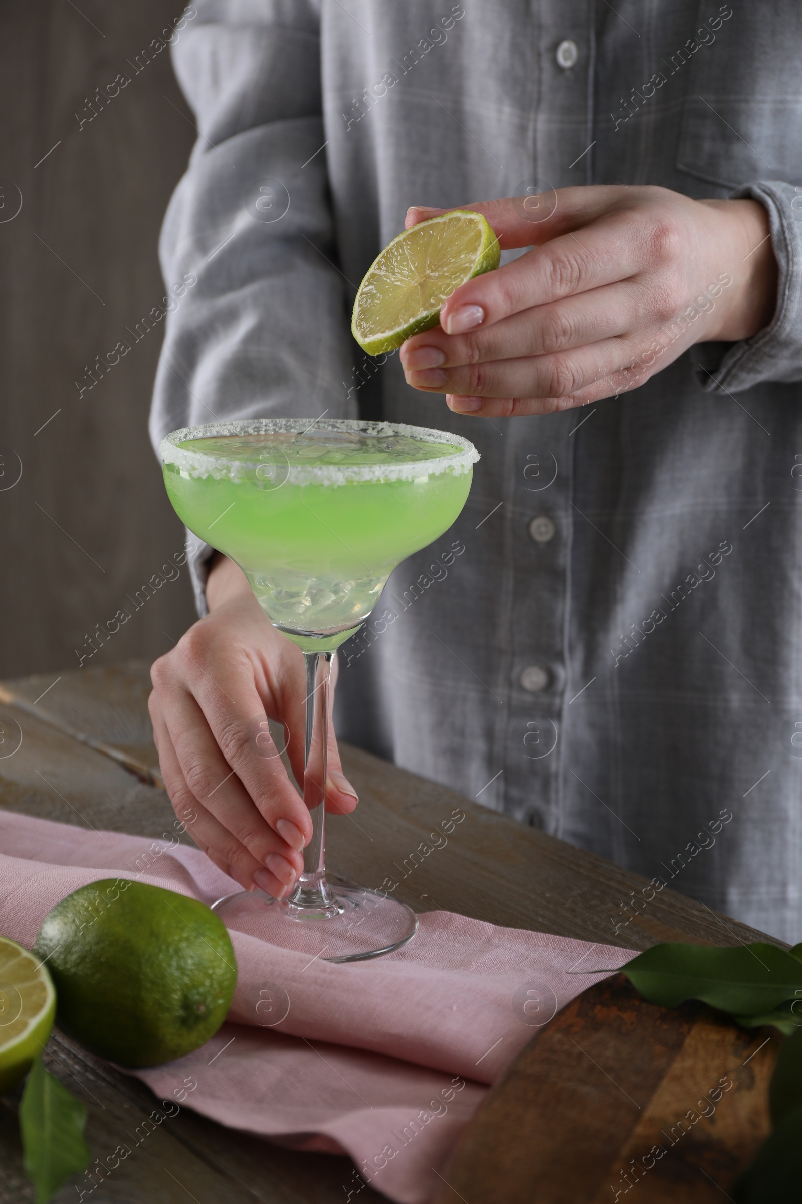 Photo of Woman squeezing lime juice into glass with delicious Margarita cocktail at wooden table, closeup