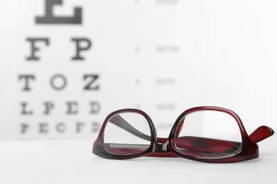 Photo of Glasses with corrective lenses on table against eye chart