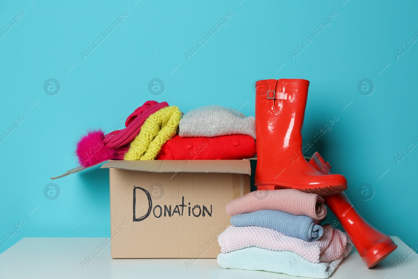 Photo of Donation box, knitted clothes and rubber boots on table against color background