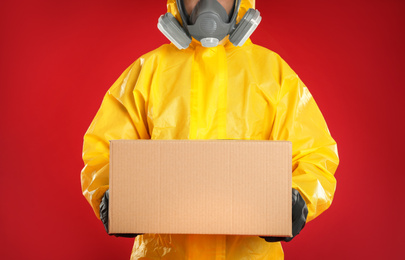 Photo of Man wearing chemical protective suit with cardboard box on red background, closeup. Prevention of virus spread