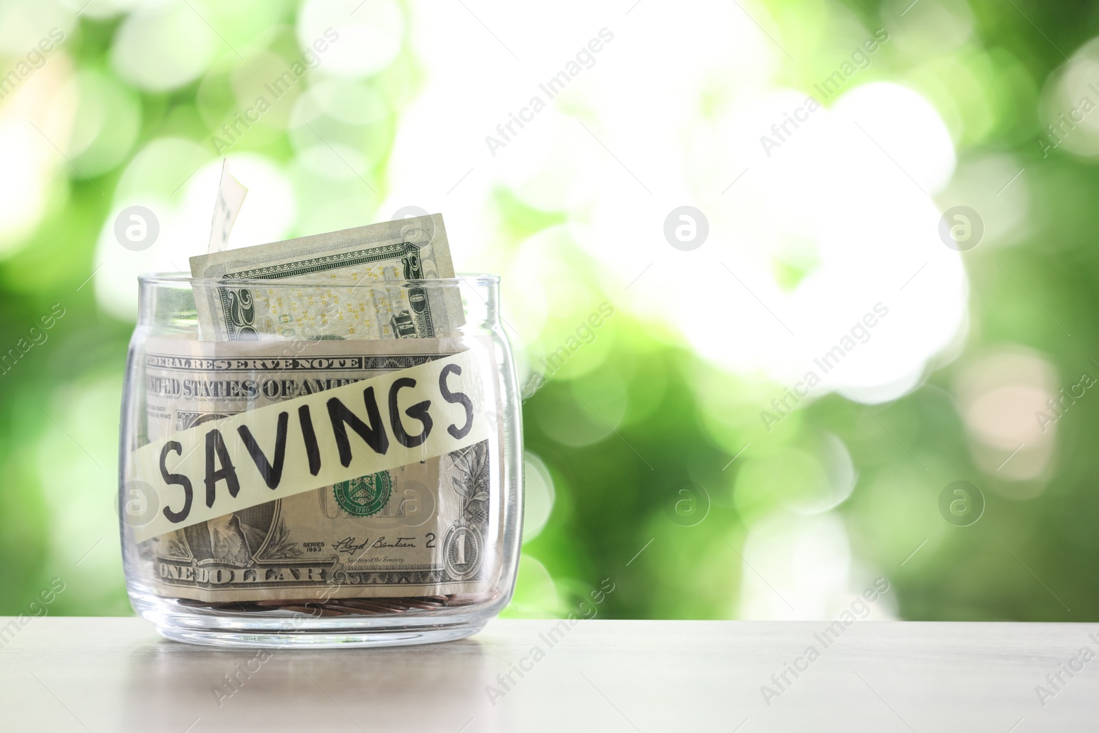 Photo of Glass jar with money and label SAVINGS on table against blurred background. Space for text