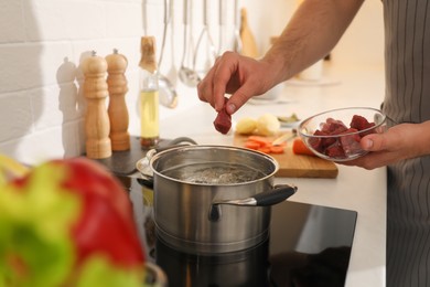 Photo of Man putting meat into pot to make bouillon in kitchen, closeup. Homemade recipe