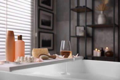 Photo of Wooden tray with wine, toiletries and flower petals on bathtub in bathroom
