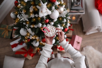 Photo of Woman with cup of delicious hot drink near Christmas tree at home, top view