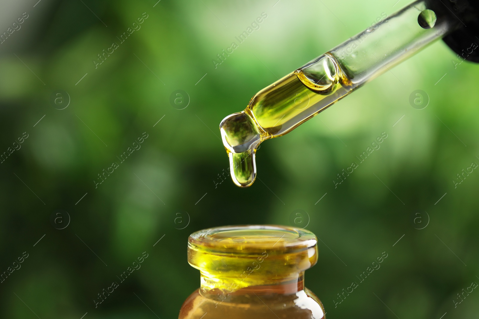 Photo of Dripping essential oil from pipette into glass bottle against blurred green background, closeup