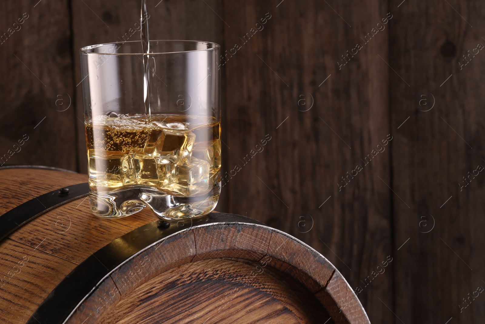 Photo of Pouring whiskey into glass on barrel against wooden background, closeup. Space for text