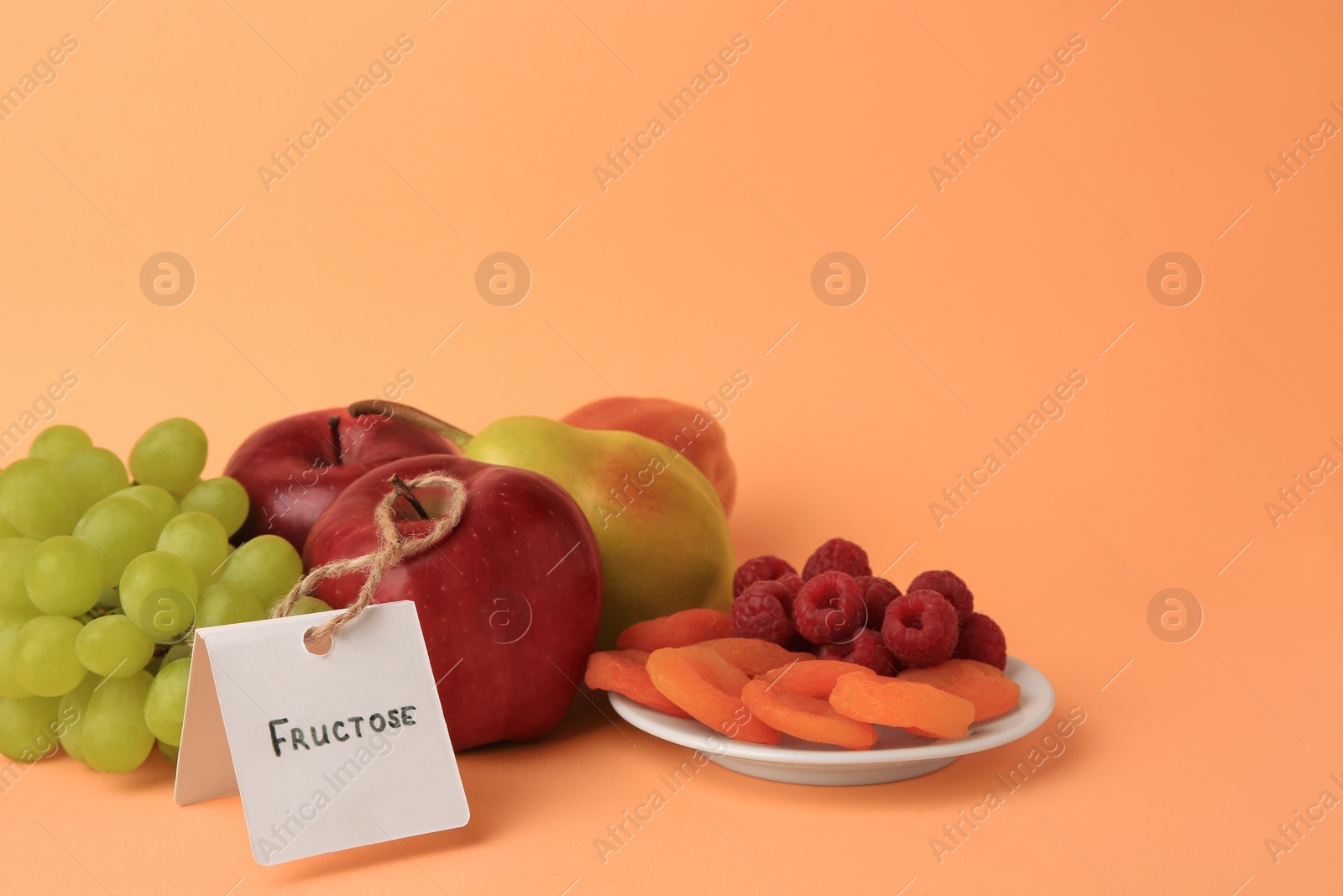 Photo of Card with word Fructose, delicious ripe fruits, raspberries and dried apricots on pale orange background. Space for text