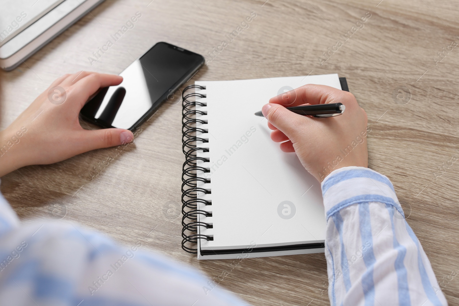 Photo of Woman with smartphone writing in notebook at wooden table, closeup