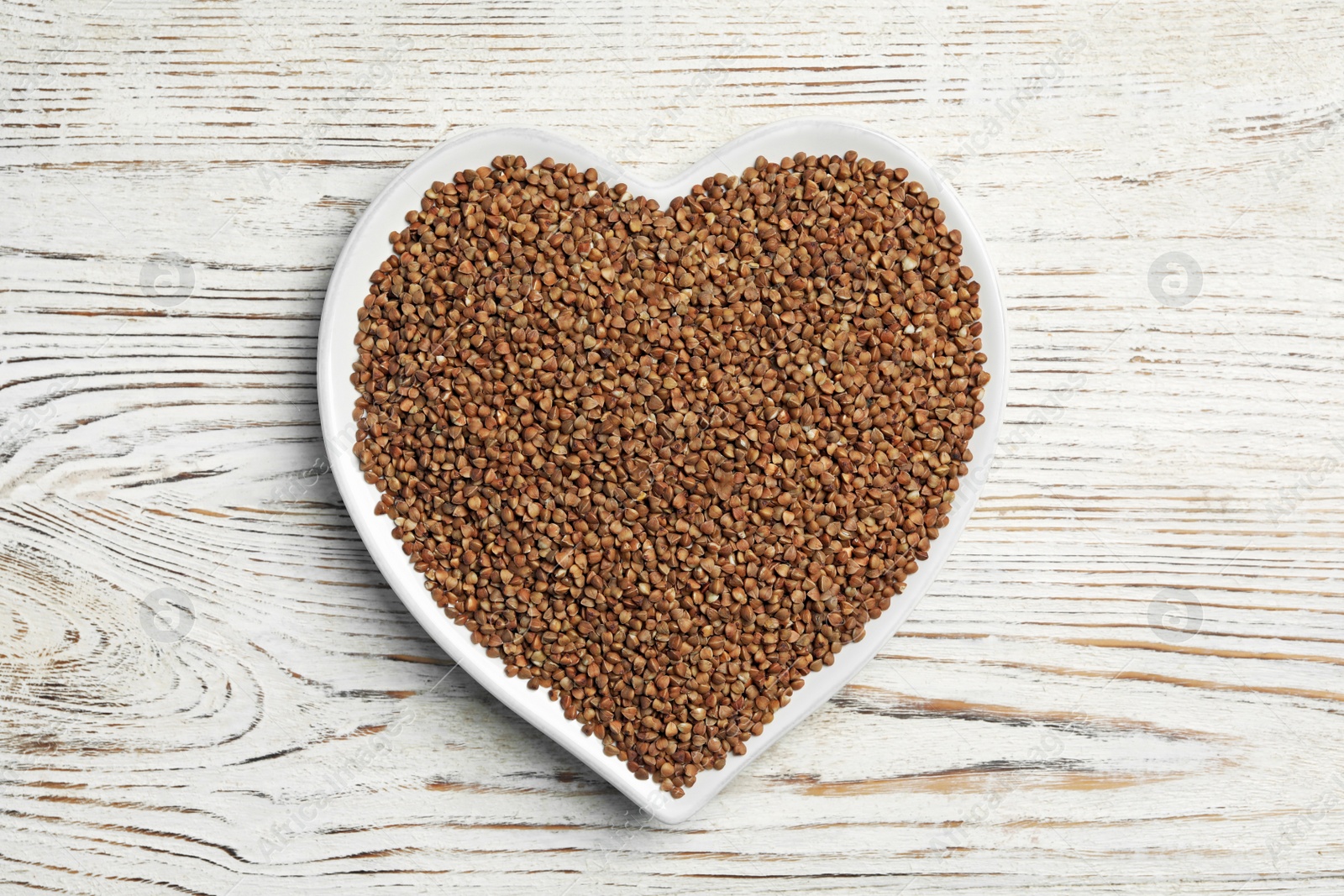 Photo of Heart shaped plate with raw buckwheat on wooden background, top view