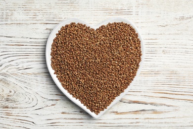 Photo of Heart shaped plate with raw buckwheat on wooden background, top view