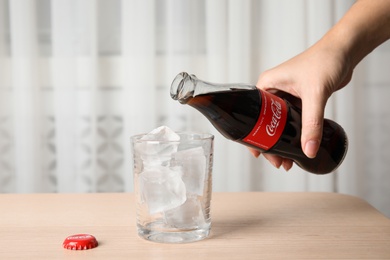 Photo of MYKOLAIV, UKRAINE - NOVEMBER 15, 2018: Woman pouring Coca Cola into glass with ice cubes at table, closeup