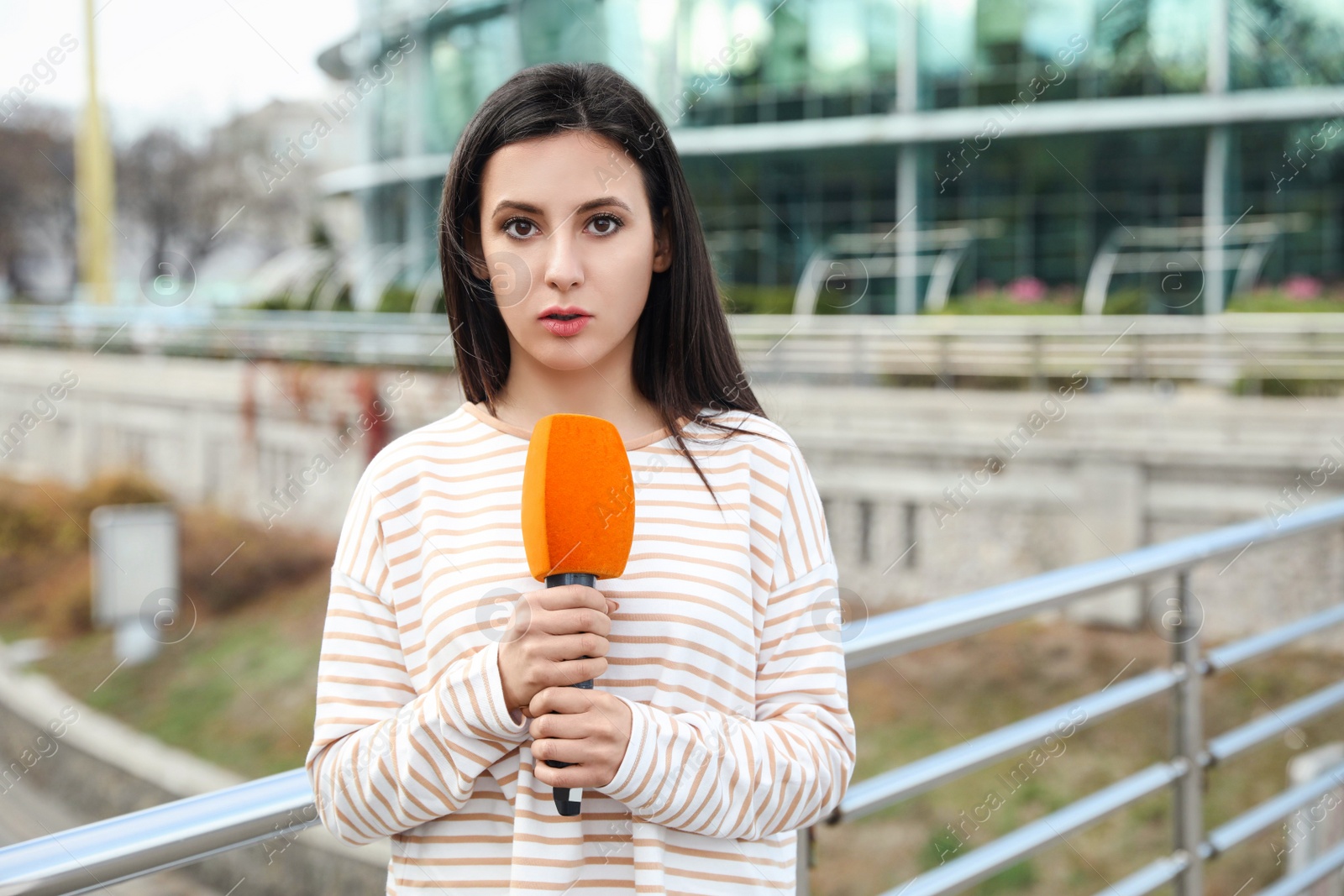 Photo of Young female journalist with microphone working on city street