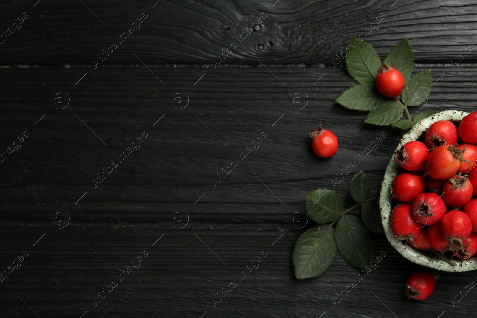Photo of Ripe rose hip berries with green leaves on black wooden table, flat lay. Space for text