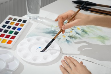 Photo of Woman painting flowers with watercolor at white table indoors, closeup. Creative artwork