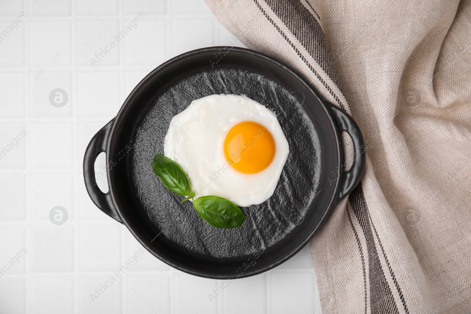 Photo of Tasty fried egg with basil in pan on white tiled table, flat lay