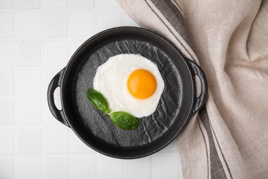 Tasty fried egg with basil in pan on white tiled table, flat lay
