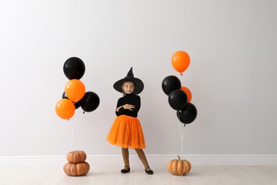 Photo of Cute little girl with balloons and pumpkins wearing Halloween costume near light wall