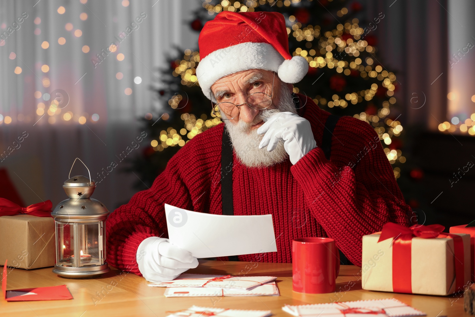 Photo of Santa Claus reading letter at his workplace in room with Christmas tree