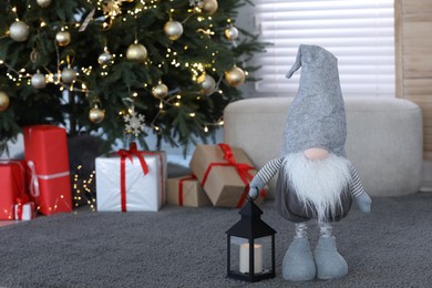 Photo of Cute Scandinavian gnome with lantern candle holder near Christmas tree on carpet in room. Space for text