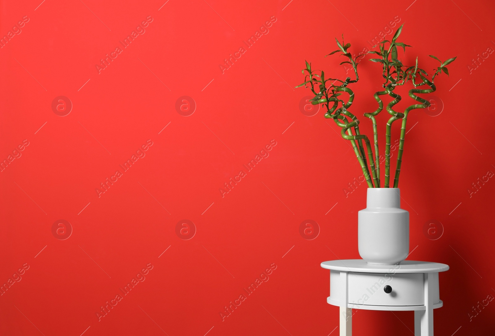 Photo of Vase with bamboo stems on white table against red background, space for text