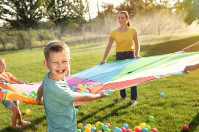 Group of children and teacher playing with rainbow playground parachute on green grass. Summer camp activity