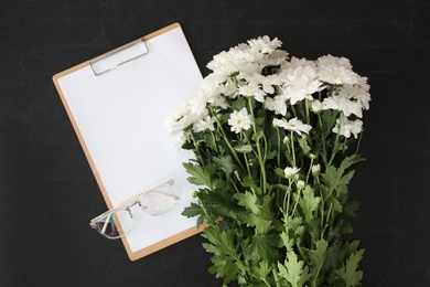 Photo of Clipboard, glasses and beautiful white flowers on blackboard, flat lay. Happy Teacher's Day