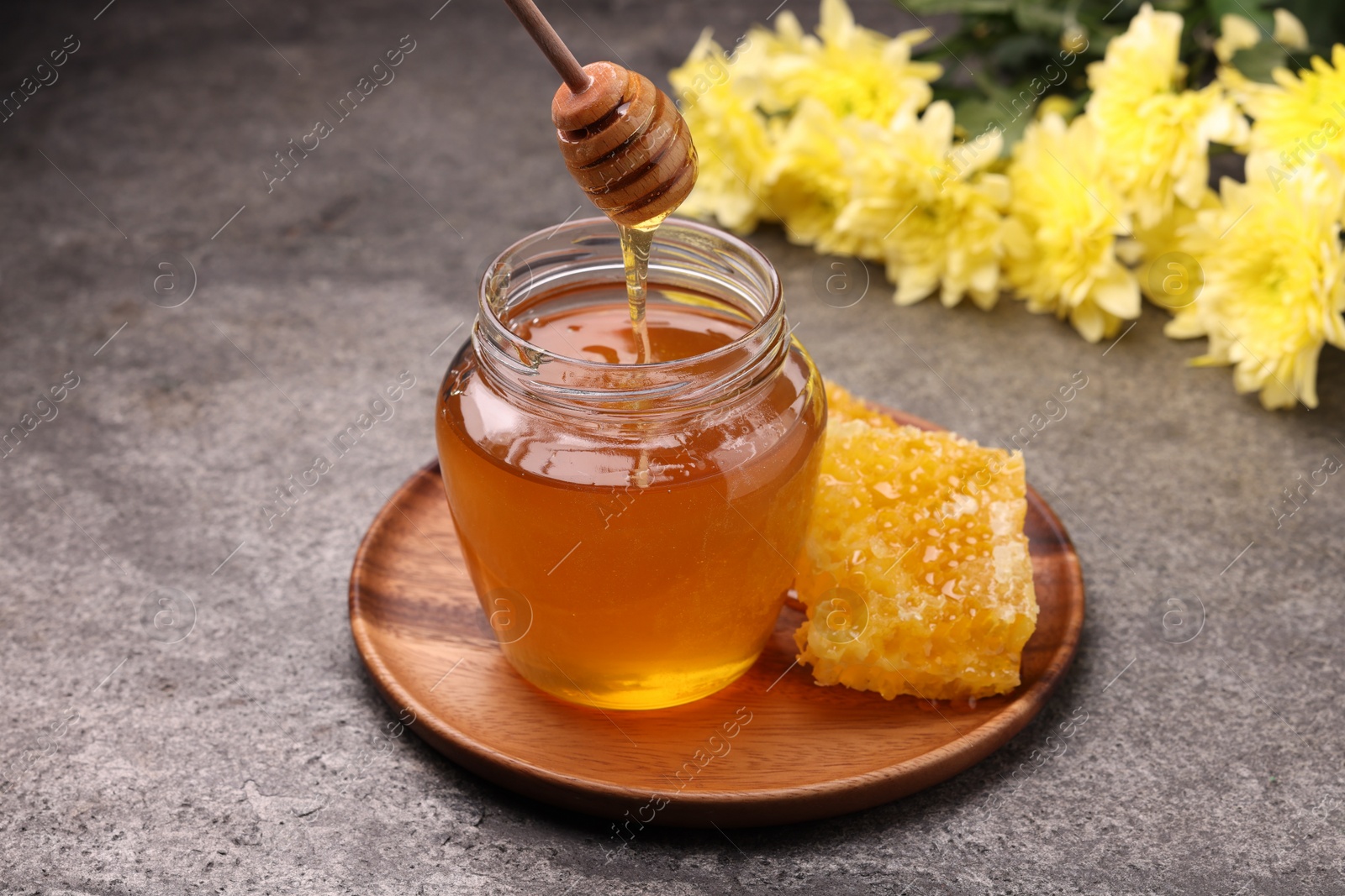 Photo of Pouring sweet golden honey from dipper into jar and honeycomb on grey textured table