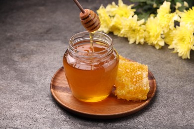 Pouring sweet golden honey from dipper into jar and honeycomb on grey textured table