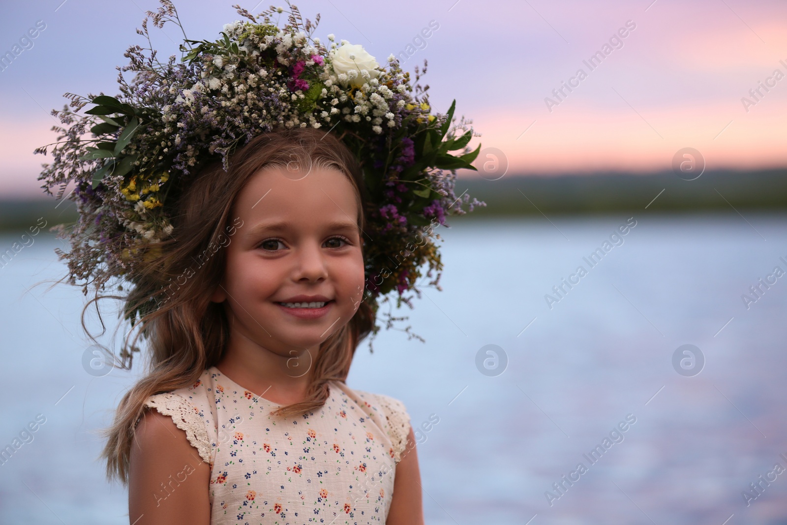 Photo of Cute little girl wearing wreath made of beautiful flowers near river at sunset
