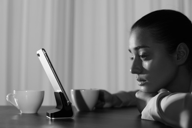 Upset woman with mobile phone at table indoors, black and white effect. Loneliness concept