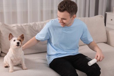 Photo of Pet shedding. Smiling man with lint roller removing dog's hair from pants on sofa at home