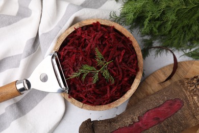 Photo of Grated red beet with dill in bowl and vegetable peeler on table, flat lay