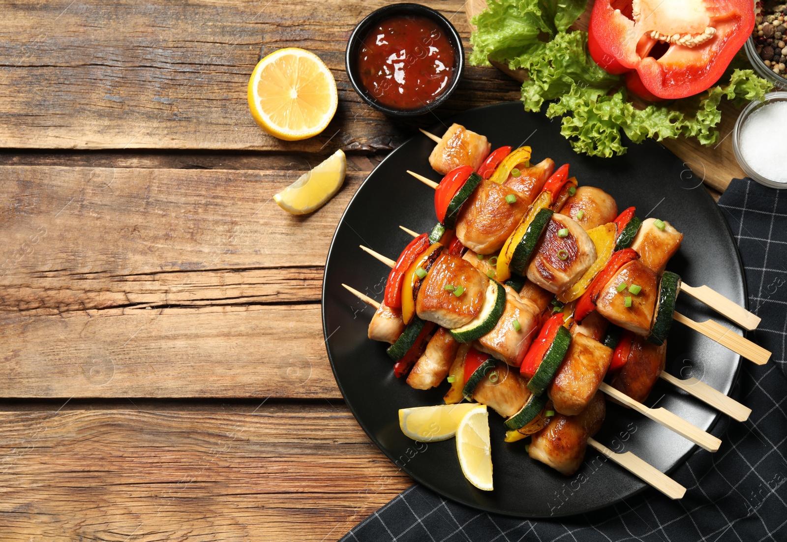 Photo of Delicious chicken shish kebabs with vegetables, ketchup and lemon on wooden table, flat lay. Space for text