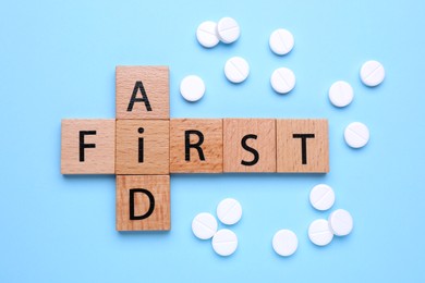 Words First Aid made of wooden cubes and pills on light blue background, flat lay