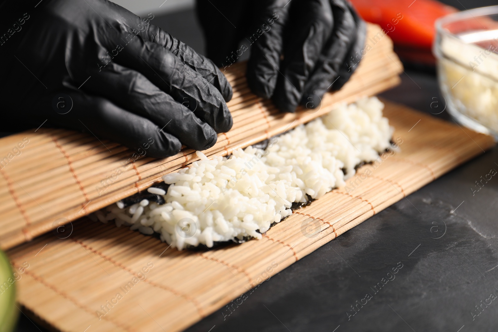 Photo of Chef in gloves wrapping sushi roll at dark textured table, closeup