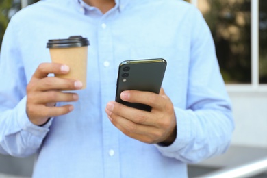 Man with cup of coffee using modern mobile phone outdoors, closeup