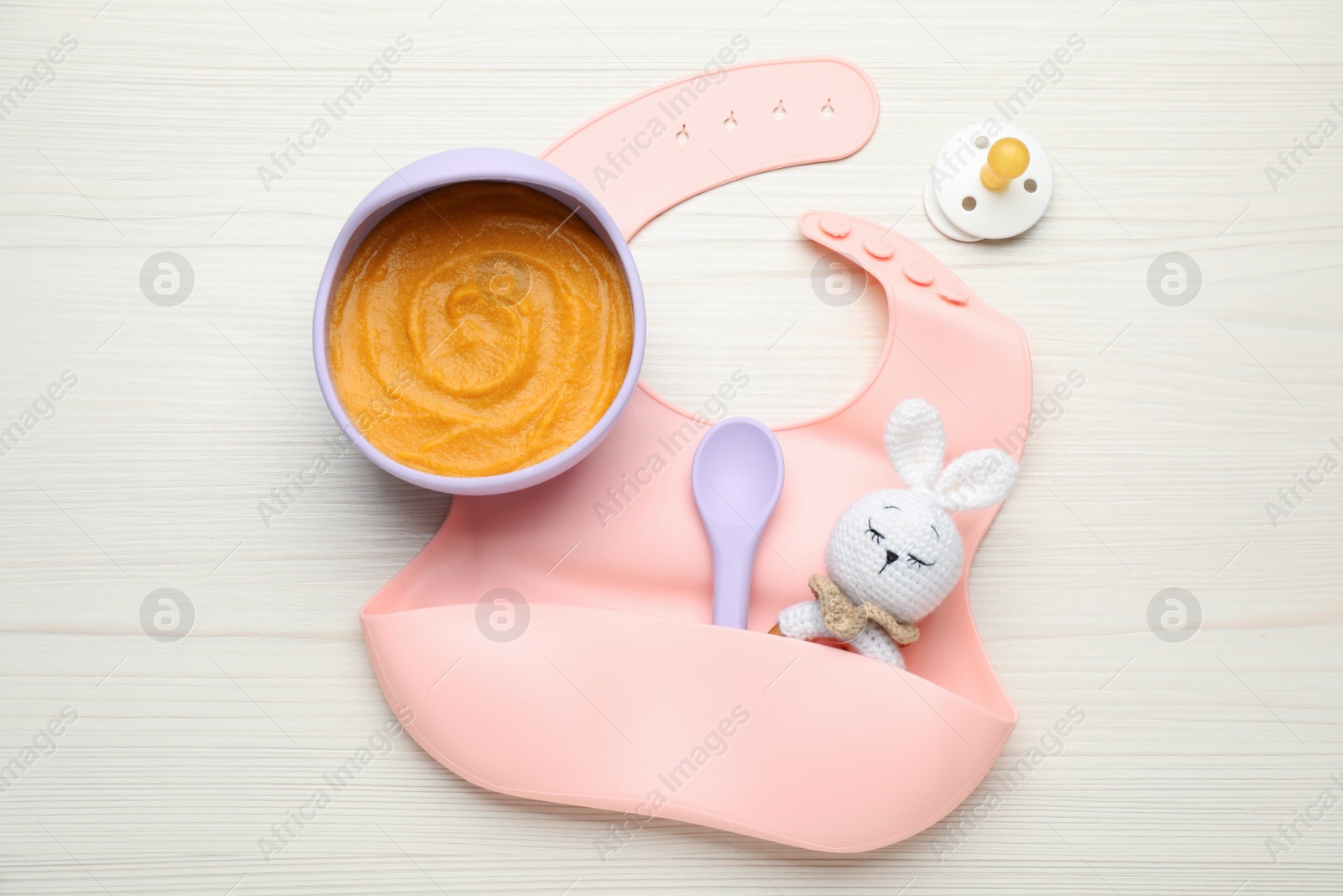 Photo of Silicone baby bib, toy and plastic dishware with healthy food on white wooden table, flat lay